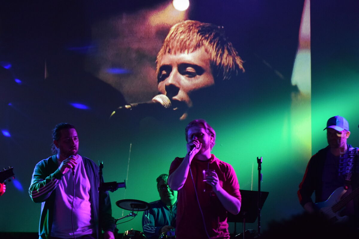 Duet performing with the band as a giant music video plays on the projection screen behind them in green and purple light