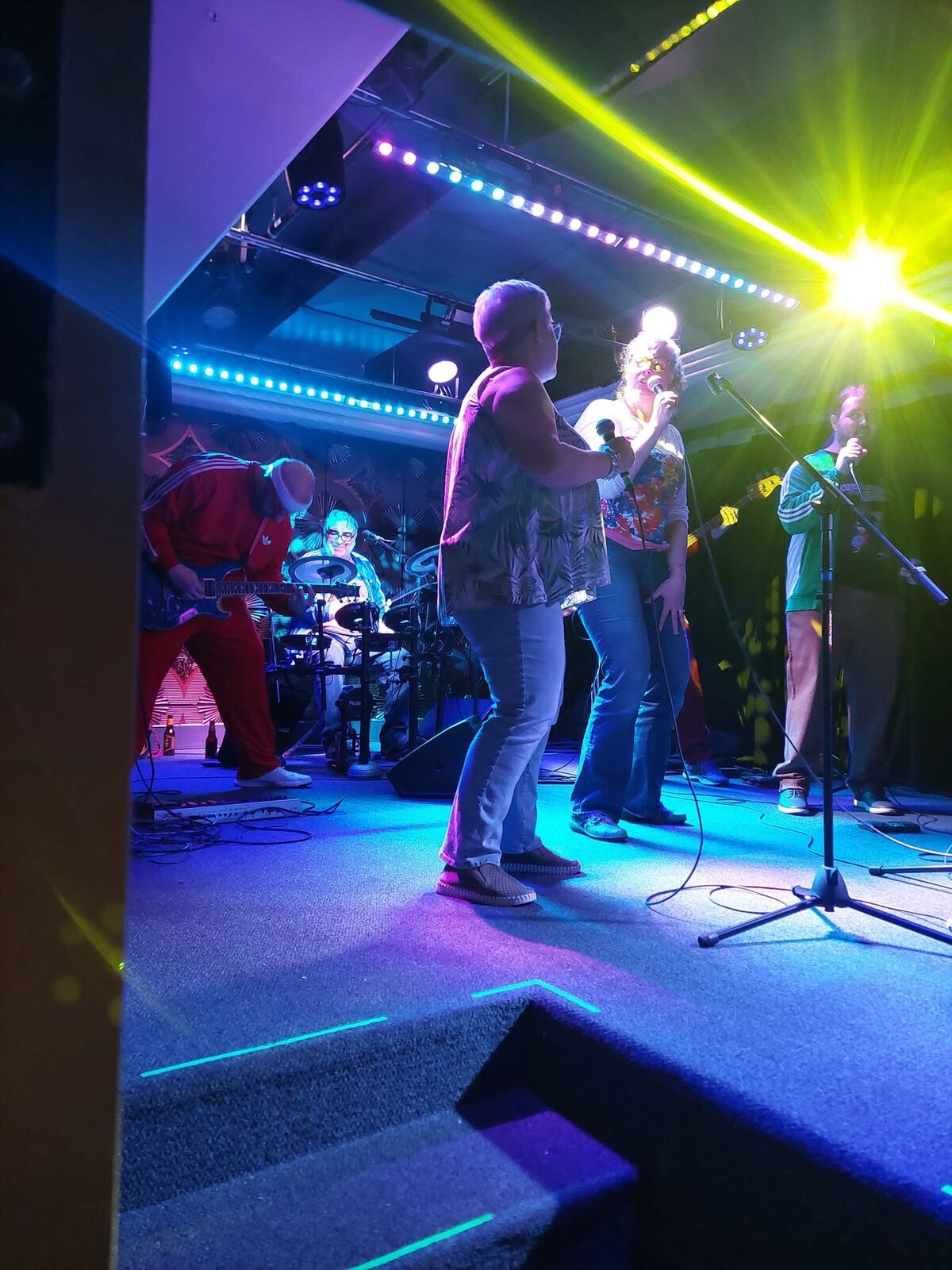 Two performers singing together on stage with the band, shot from a low angle with the lighting rig above